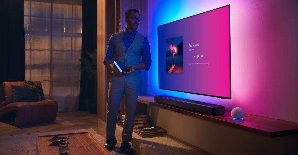 Müzik arayüzü ve Fidelio ses çubuğu bulunan Philips Ambilight TV'nin yakınındaki modern bir oturma odasındaki kişi