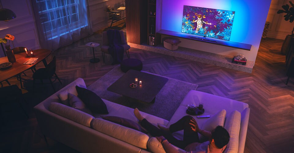 Loş ışıklı bir oturma odasındaki kanepede dinlenen ve Philips Ambilight TV izleyen adam