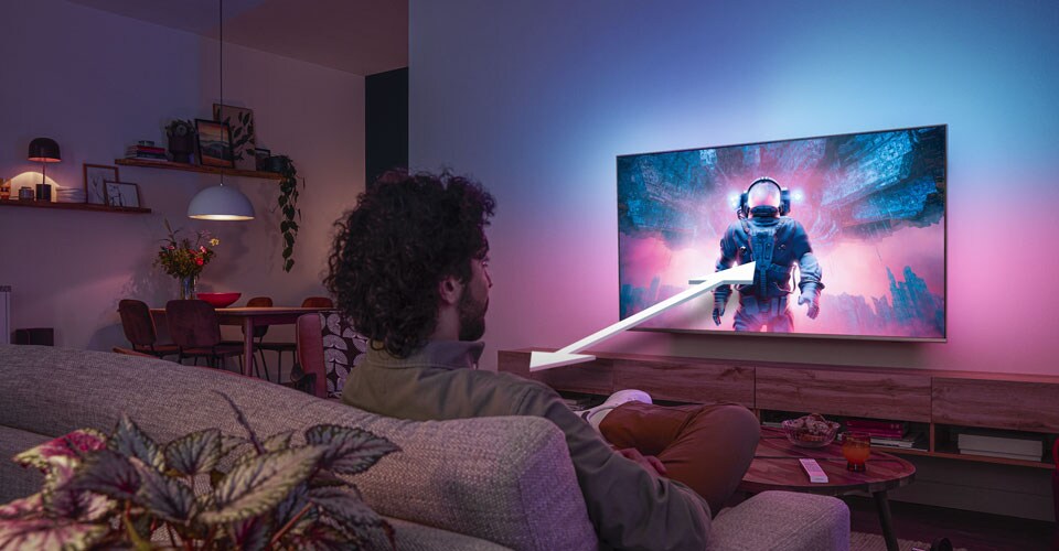 Renkli bir astronot sahnesi gösteren Philips Ambilight TV'yi izleyen ve optimum izleme mesafesini gösteren bir ok bulunan bir kanepede oturan adam